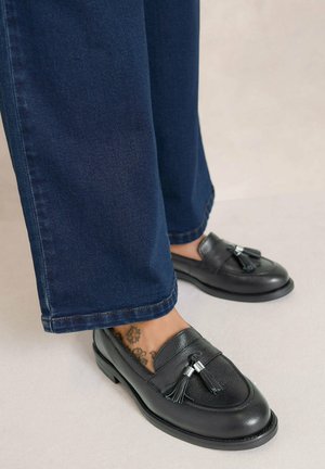 Mocassins à glands en cuir noir portés avec un jean droit bleu foncé sur un sol clair, tatouages visibles aux chevilles.