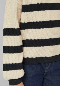 Sweter w paski w beżowym i czarnym kolorze; posiada szerokie, poziome paski. Teksturowana tkanina z ściągaczami przy mankietach i dole. Połączony z niebieskimi dżinsami.