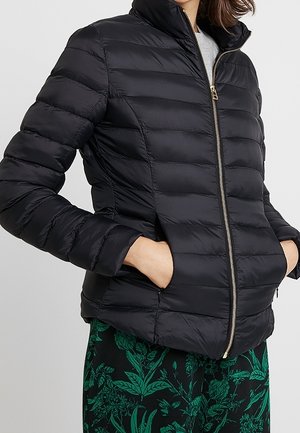 Femme portant une veste matelassée noire zippée avec les mains dans les poches, associée à un pantalon vert à motif floral.