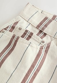 Gros plan sur un tissu crème à rayures verticales marron et noir, avec une ceinture et une poche fixée par des rivets métalliques.