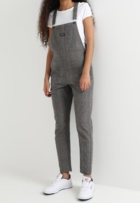 Graue Hahnentrittmuster-Overalls mit verstellbaren Trägern, enganliegenden Beinen und einer Fronttasche. Tragen über einem weißen T-Shirt und kombiniert mit weißen Sneakern.