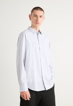 Light blue button-up shirt with vertical stripes, featuring embroidered "Calvin Klein" logo on the chest, long sleeves, and curved hem.