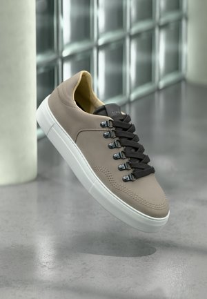 Sneaker beige con lacci neri e suola bianca che sembra fluttuare sopra un pavimento in cemento lucido, con una parete di blocchi di vetro e una colonna sullo sfondo.
