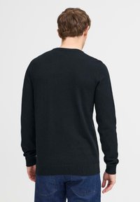 Uomo con capelli corti castani che indossa un maglione nero a maniche lunghe e jeans blu, visto da dietro su uno sfondo uniforme.