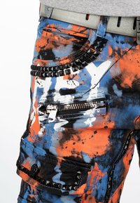 Shorts cargo multicolores avec un design abstrait de taches de peinture en bleu, orange et noir, ornés de zips et d'accents cloutés.