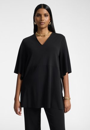 Femme aux longs cheveux noirs portant un haut noir ample à col en V, un pantalon noir, un collier chaîne doré, des boucles d'oreilles et un bracelet, debout sur un fond neutre.