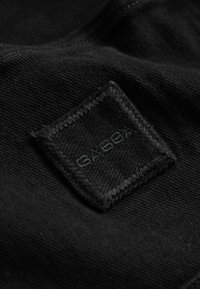 Black fabric with a stitched square patch featuring the embossed brand name "G-STAR" in matching black thread.
