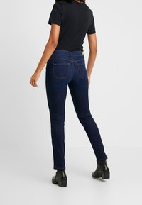 Mörkblå skinny jeans i denim med en medelhög midja och två bakfickor med guldtrådsdetaljer. Bärs tillsammans med en svart t-shirt.