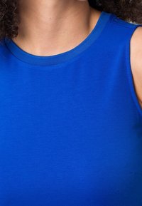 Femme portant un haut bleu sans manches avec un col rond, court au niveau du cou et des épaules.
