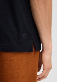 Schwarz kurzärmliges Shirt mit strukturiertem Finish, ausgestattet mit seitlichen Schlitzen und einem dezenten Logo am Saum. Kombiniert mit orangefarbenen, strukturierten Hosen.