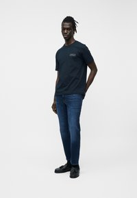 Férfi áll fekete Tommy Hilfiger pólóban, sötétkék farmernadrágban és fekete mokaszinban, egyszerű fehér háttér előtt.