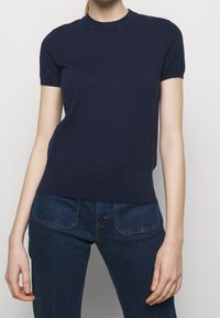 Mujer vistiendo un suéter azul marino ajustado de manga corta y jeans de mezclilla azul oscuro, de pie contra un fondo claro y liso.