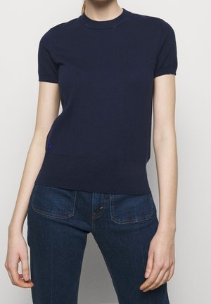 Kvinde iført en tætsiddende navyblå kortærmet sweater og mørkeblå denimjeans, der står foran en ensfarvet lys baggrund.