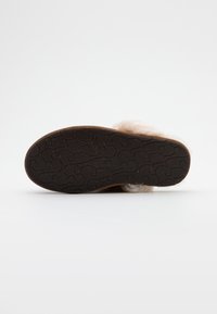 Vue unique de la semelle d'une pantoufle douce beige avec une semelle en caoutchouc brun foncé présentant un motif répétitif en forme de "S".