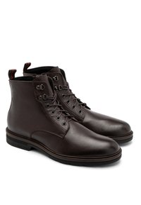 Brązowe skórzane botki za kostkę o gładkiej fakturze, okrągłym nosku, systemie sznurowania oraz pętlach do zakładania z czerwonymi akcentami z tyłu.