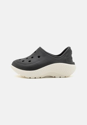 Musta slip-on-puha, jossa on päällä ilmanvaihtoaukot ja paksu valkoinen kuvioitu pohja, suunniteltu mukavuutta ja rentoa käyttöä varten.