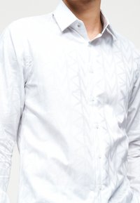 Άνδρας που φοράει λευκό πουκάμισο με κουμπιά, με διακριτικό σχέδιο σε σχήμα κεραυνών και μυτερό γιακά, εμφανίζεται από τη μέση μέχρι τον λαιμό.