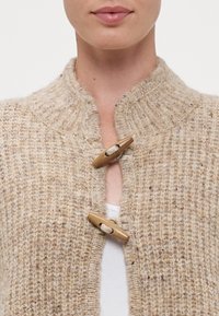 Garnitör i beige ullblandning, med en ribbad krage och två träknappar vid framöppningen. Texturen verkar varm och mysig.