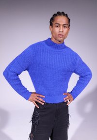 Un pull bleu épais tricoté avec un col montant et une texture côtelée, associé à un jean noir déchiré. Matériau doux et duveteux.