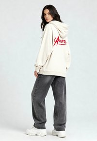 Kreemist kapuutsiga pusa, millel on seljal punane "Amare" tekst, koos lahti istuvate tumedate hallide teksapükstega ja valgete tossudega. Pehme kangas, vabalt langev disain.