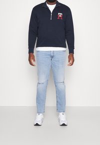 Marinblå zip-up sweatshirt med röd och vit broderad logotyp, i kombination med ljusblå slitna jeans och vita sneakers. Enkelt, avslappnat outfit.