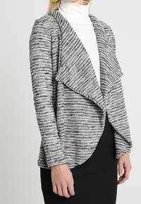 Open-front cardigan made of textured, black-and-white striped knit material; features draped collar and sleek silhouette, paired with a white turtleneck.