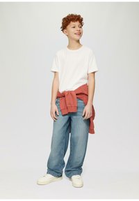 Witte t-shirt, losse blauwe jeans en een roestkleurige sweater om de taille geknoopt. Witte sneakers maken de outfit compleet.