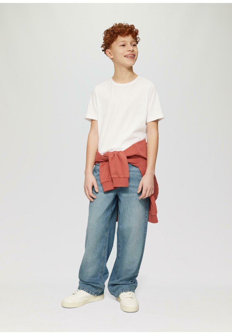 Witte t-shirt, losse blauwe jeans en een roestkleurige sweater om de taille geknoopt. Witte sneakers maken de outfit compleet.