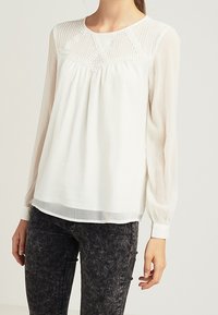 Bílá halenka s dlouhým rukávem, s texturovaným průhledným vrškem a krajkovými detaily, s volným střihem a kulatým výstřihem, doplněná o tmavé džíny.