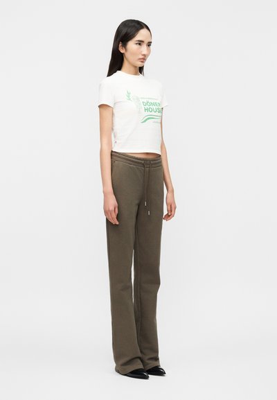 Νεαρή γυναίκα που στέκεται, φοράει λευκό κοντό T-shirt με πράσινο κείμενο "Döner House" και χακί παντελόνια με κορδόνι στη μέση και μαύρα παπούτσια.