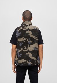 Fleeceväst med hög krage i kamomönster, med jordnära toner av svart, grön och blå. Mjuk, texturerad utsida synlig från baksidan.