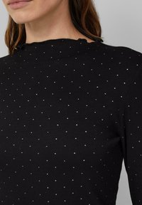 Zwarte geribbelde top met kleine zilveren stippen, een textuurstof en een golvende halslijn. Tailored fit met lange mouwen.