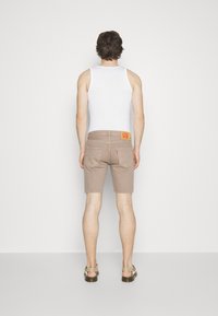 Pantalones cortos de mezclilla beige con un corte recto, que presentan bolsillos traseros y una etiqueta cosida en la cintura, combinados con una camiseta sin mangas blanca.