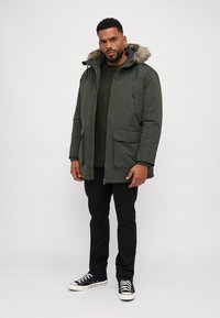 Parka de color verde oliva con capucha con borde de piel, cierre de cremallera y botones, dos grandes bolsillos frontales, llevada sobre un suéter verde oscuro y pantalones negros.