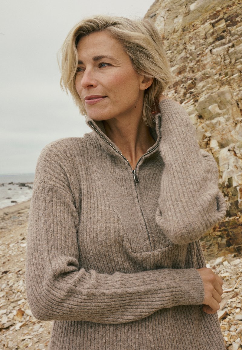 Pull en maille côtelée beige avec col zippé et détails torsadés sur les manches, doté d'un tissu doux et texturé.