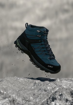 Une chaussure de randonnée CMP turquoise et noire flotte au-dessus d’un sentier rocailleux, tandis que de petits cailloux se dispersent en dessous.