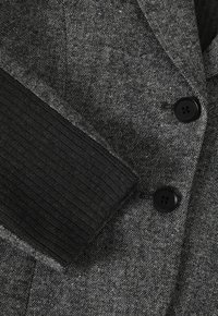 Šedý blazer s texturovanou tkaninou, vyznačující se jemným herringbone vzorem, černými žebrovanými manžetami a dvěma černými knoflíky vpředu.