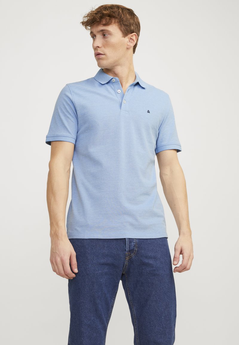 Hellblaues Poloshirt mit strukturiertem Finish, drei Knöpfen am Kragen und einem kleinen schwarzen Logo auf der Brust. Kombiniert mit dunklen Jeans.