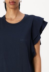 Tumesinine lühikeste varrukatega särk, millel on rippuvad varrukad, ümar kaelus ja reljeefne "LIU-JO MILANO" logo ees. Sile puuvillane kangas.