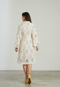Robe bianca con intricate decorazioni a diamante, maniche lunghe e vita asciugata. Il tessuto textured aggiunge dimensione. Indossata con tacchi a cuneo beige.