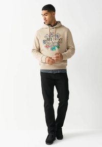 Beige hoodie med et trykt grafisk logo med farverige designs, parret med sorte bukser og sorte sko, sat mod en neutral baggrund.