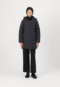 Zwarte pufferjack met een chevronpatroon, ritssluiting en capuchon. Draagt het over een zwart top, gecombineerd met brede zwarte broek, staan op een effen achtergrond.
