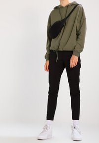 Felpa cropped verde oliva, pantaloni neri slim-fit, sneakers bianchi e una borsa a tracolla nera. Presenta un orlo con coulisse e una texture morbida.
