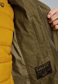 Intérieur d'une veste vert olive avec une couche matelassée jaune. Comprend une fermeture éclair en métal et une étiquette noire avec du texte blanc sur le côté gauche.