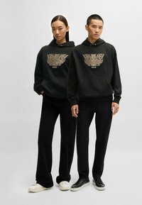 Svarta huvtröjor har en gyllene örn-grafik och "BOSS"-logotyp. Modellerna bär svarta byxor och sneakers, en med vita sulor och en med svarta.