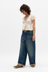 Kvinne iført kremfarget kortermet V-hals topp med blonder, løse vidde blå jeans med oppbrett på kanten, og olivengrønne sko som står på hvit bakgrunn.