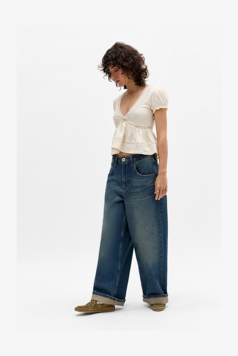 Kvinne iført kremfarget kortermet V-hals topp med blonder, løse vidde blå jeans med oppbrett på kanten, og olivengrønne sko som står på hvit bakgrunn.