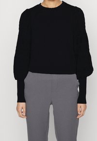 Person i en svart långärmad blus med texturerade puffärmar och grå högmidjade byxor, stående med avslappnade armar.