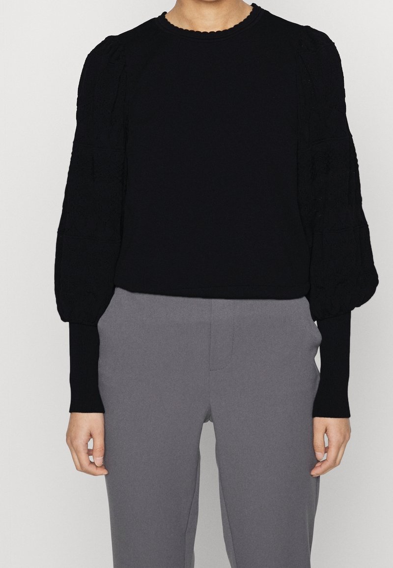 Person i en svart långärmad blus med texturerade puffärmar och grå högmidjade byxor, stående med avslappnade armar.