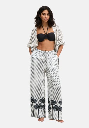 Frau trägt ein schwarzes Bandeau-Top mit einem weißen, gemusterten offenen Hemd und weit geschnittene Hosen mit schwarzen Palmenmotiven am Saum.